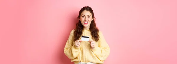 兴奋的女孩拿着塑料信用卡出去购物 对着相机笑着 站在粉色的背景下 — 图库照片