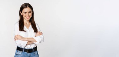 Smiling young businesswoman, female entrepreneur in white shirt, cross arms on chest like professional, standing over studio background.