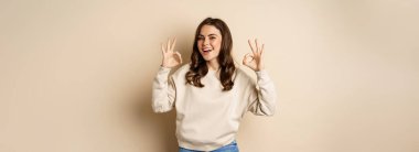 Very good, nice work. Cheerful young woman agrees, shows okay, ok praise, recommending smth, standing over beige background.