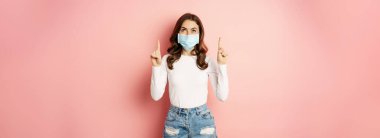 Portrait of young glamour girl in medical face mask, reading promo text, pointing and looking up, standing over pink background. Covid and pandemic concept