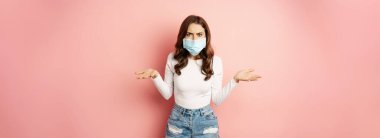 Covid-19. Confused young woman in medical face mask, shrugging shoulders and looking clueless, standing puzzled against pink background.