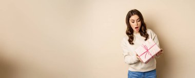 Excited beautiful girl with pink wrapped gift box, receive presents, standing over beige background.