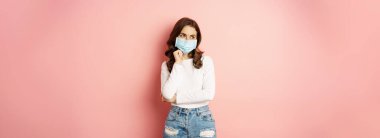 Portrait of stylish modern girl in medical face mask, looking coquettish and smiling at camera, standing over pink background. Pandemic and covid concept.