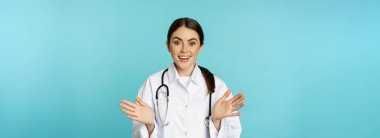 Happy and excited smiling young woman looking with excitement and surprised, standing in white lab coat and stethoscope, blue background.