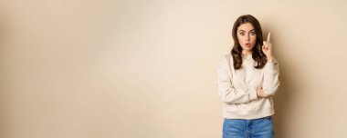 Young woman suggests an idea, raising finger, has revelation, pitching a plan, pointing up, standing over beige background.