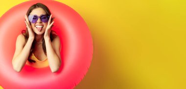Close up portrait of enthusiastic young woman inside pink swimming ring, laughing and smiling, enjoying beach holiday, summer vacation, yellow background.