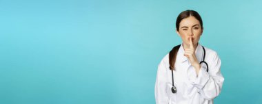 Smiling cute doctor woman, shush, press hush shh sign with finger pressed to lips, hiding secret, taboo gesture, standing over torquoise background.
