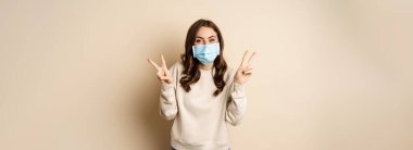 Covid-19, pandemic and quarantine concept. Beautiful adult woman in face medical mask, showing peace, v-sign gesture, standing over beige background.