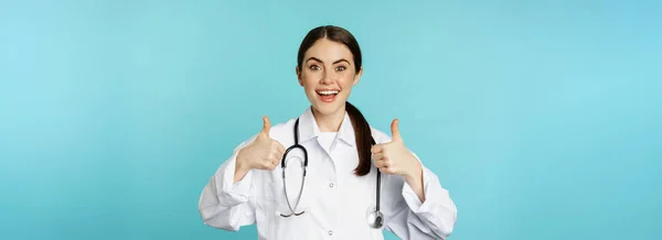 青い背景の上に立って賞賛のような承認で親指を示す白いコートの幸せな女性医師 医療従事者 — ストック写真