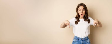 Surprised modern womn pointing fingers down, gasping amazed, showing smth awesome below, standing over beige background.