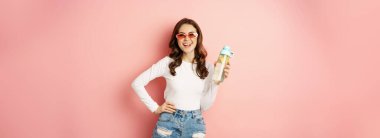 Stylish girl in spring outfit, wearing sunglasses, holding water bottle with lemon, healthy drink, laughing and smiling, standing over pink background.