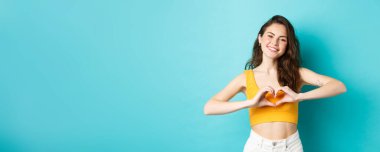 Yaz tatili ve duygu konsepti. Genç romantik kız seni sevdiğimi gösteriyor. Kalp hareketi, gülümseme, mavi arka planda durmak..