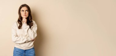 Image of beautiful woman with serious face thinking, standing over beige background.