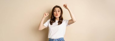 Enthusiastic adn surprised woman winning, dancing and celebrating, standing in tshirt over beige background.