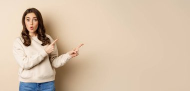 Surprised brunette woman pointing fingers right, showing advertisement and say wow impressed, standing over beige background.