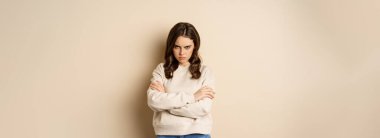Angry girlfriend sulking, cross arms on chest and look offended, staring moody at camera, standing over beige background.