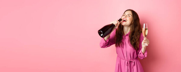 Celebration and holidays concept. Happy carefree woman drinking champagne from bottle with pleased smile, relaxing on party, holding glass, standing over pink background.