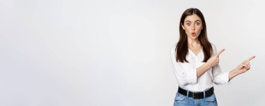 Amazed young woman pointing fingers down, showing announcement or logo banner, looking surprised and intrigued, standing over white background.