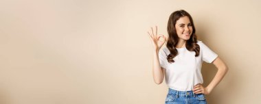 Stylish young woman smiling satisfied, showing okay, ok sign in approval, recommending, standing over beige background.