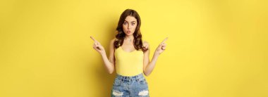 Portrait of stylish young female model, showing advertisement, pointing at logo or banner, demonstrating two ways, choices in store, standing over yellow background.