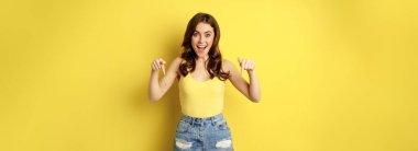 Look down, big news. Portrait of amazed young woman pointing down below, smiling excited, showing logo or banner, standing over yellow background.