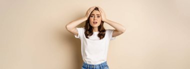 Young woman facing disaster, looking anxious in panic, holding hands on head frustrated, standing over beige background.