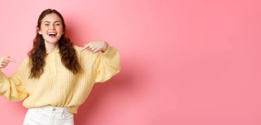 Kendinden emin mutlu kız kendini gösteriyor ve gülüyor, kendini tanıtıyor ve gülümsüyor, pembe arka plana karşı duruyor..