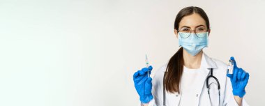 Covid-19 vaccination and healthcare concept. Young doctor in medical mask showing syringe and vaccine from coronavirus omicron variant, standing over white background.