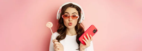 身披棒棒糖和手机 戴着太阳镜和耳机 听着音乐 站在粉色背景上的时髦年轻女子 — 图库照片