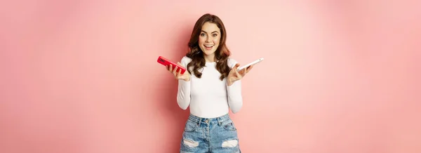 stock image Technology, spring promo concept. Stylish glamour woman holding two smartphones, mobile phones in both hands, smiling pleased, buying new cellphone, pink background.
