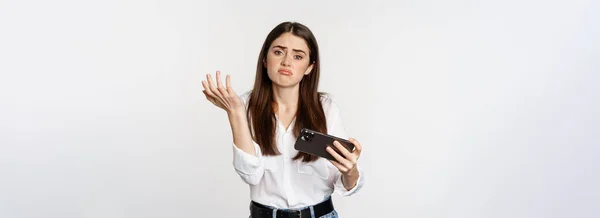 悲しい女性は モバイルビデオゲームで失わ スマートフォンで動揺と失望を見て 説得力のある 白い背景の上に立って — ストック写真
