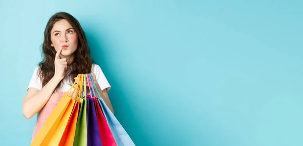 头脑清醒的女人拿着购物袋 一边摸着脸 一边回头看 一边想 在商店里做出选择 站在蓝色的背景上 — 图库照片