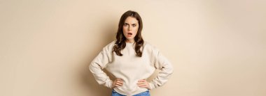 Frustrated, offended young woman looking hurt ad insulted at camera, standing shocked over beige background. People and emotions concept