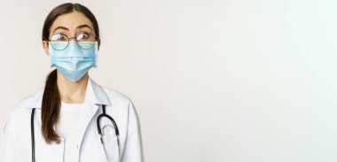 Tıp çalışanı, genç doktor şaşırmış ve şaşırmış görünüyor, yüz maskesi takıyor, beyaz arka planda dikiliyor..