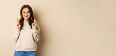 Confident woman praying, making wish with fingers crossed for good luck, standing over beige background.