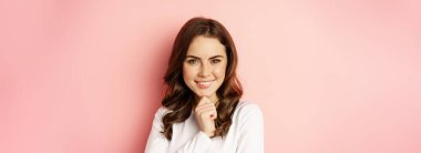 Close up portrait of thinking girl, smiling and looking confident, thoughtful face expression, standing over pink background.