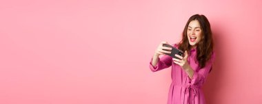 Teknoloji konsepti. Kaygısız genç bir kadın, akıllı telefonu yatay olarak tutuyor, telefonda video oyunu oynuyor, pembe arka planda duruyor. Boşluğu kopyala