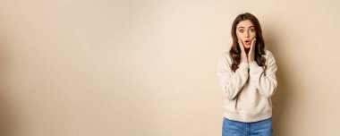 Image of surprised, amazed brunette girl gasping, looking fascinated at camera, standing over beige background.