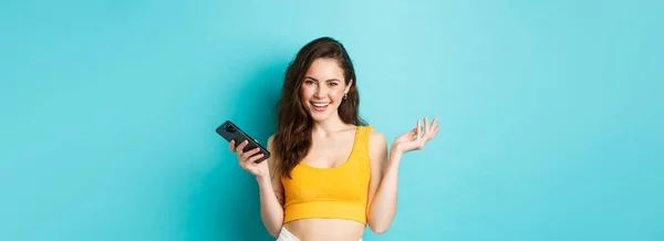时尚快乐的女人拿着智能手机 自信地微笑着 用手机聊天 站在蓝色的背景上 — 图库照片