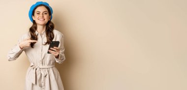 Portrait of stylish modern woman in outerwear, pointing at mobile phone screen and looking happy, smiling pleased, standing over beige background.