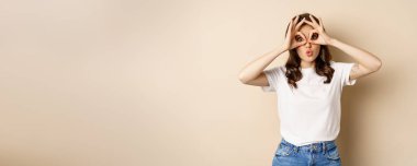 Funny brunette woman having fun, showing finger glasses gesture and fool around, posing over beige background.