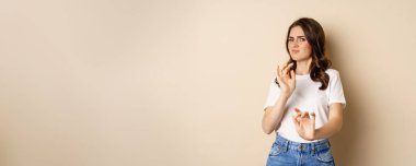Young woman cringe, shaking hands to decline, reject disgusting bad offer, standing over beige background. Copy space