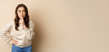 Happy coquettish girl say shh, shushing, keep secret, be quiet gesture, smiling at camera, standing over beige background.