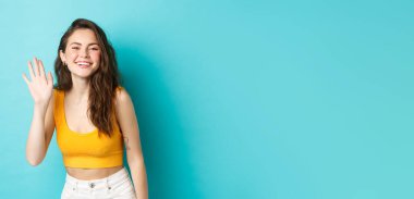 Yaz tatili ve duygu konsepti. Mükemmel vücutlu güzel, dost canlısı bir kadın, el sallıyor ve merhaba diyor, selamlıyor, mavi arka planda duruyor..