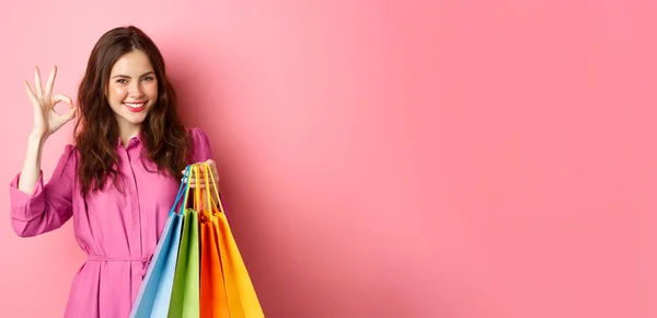 年轻快乐的女购物者展示出不错的标志 对打折很满意 买了便宜货 拿着购物袋 开心地微笑着 粉红的背景 — 图库照片