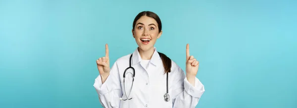 熱狂的な医療従事者 白いコートの若い女性医師 聴診器 広告を表示し 指を指して ターコイズの背景の上に立って — ストック写真