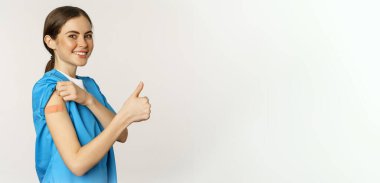 Smiling nurse, doctor in scrubs, medical worker showing her vaccinated shoulder with patch, thumbs up, recommending vaccinate from coronavirus, white background.