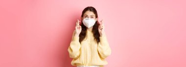 Covid-19, tecrit ve salgın konsepti. Genç bir kadın umut dolu görünüyor, şans diliyor, tıbbi maske takıyor, Coronavirus solunum cihazı takıyor, pembe arka plan..