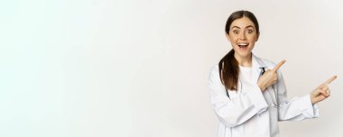 Woman doctor, physician in medical uniform, pointing fingers right, showing banner advertisement, smiling amazed, checking out promo discount, white background.