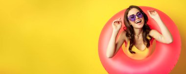 Beautiful happy woman enjoying summer vacation, wearing pink swimming ring on beach, laughing and smiling, standing over yellow background.